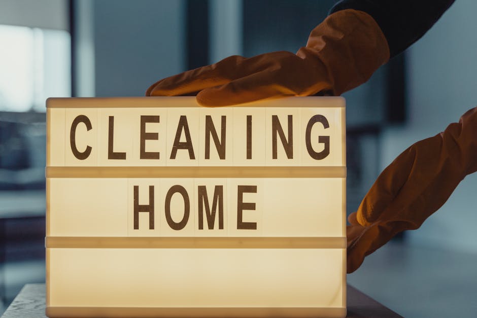 A person wearing yellow rubber gloves is holding a lightbox with the words 'CLEANING HOME' in bold black letters. The lightbox is placed on a surface in a well-lit room, emphasizing the theme of domes