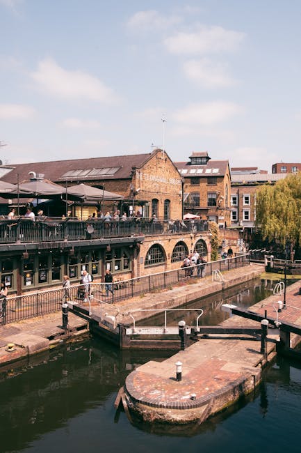 Exterior view of Camden Market showcasing brick buildings with arched windows and outdoor seating areas on a balcony. The scene includes a canal with a small dock and bollards in the foreground, along