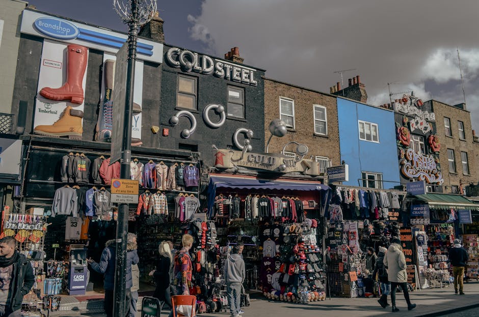 Exterior view of Camden Market showcasing brick buildings with arched windows and outdoor seating areas on a balcony. The scene includes a canal with a small dock and bollards in the foreground, along