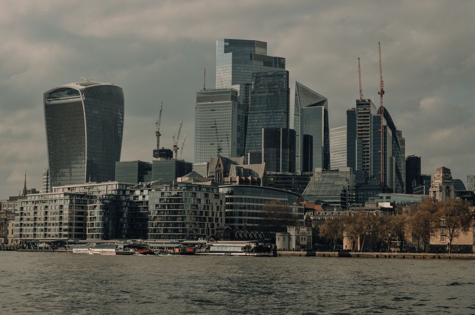 A panoramic view of Canary Wharf in London displaying modern office buildings with glass facades, including a mix of tall skyscrapers and mid-rise structures, some under construction with cranes visib