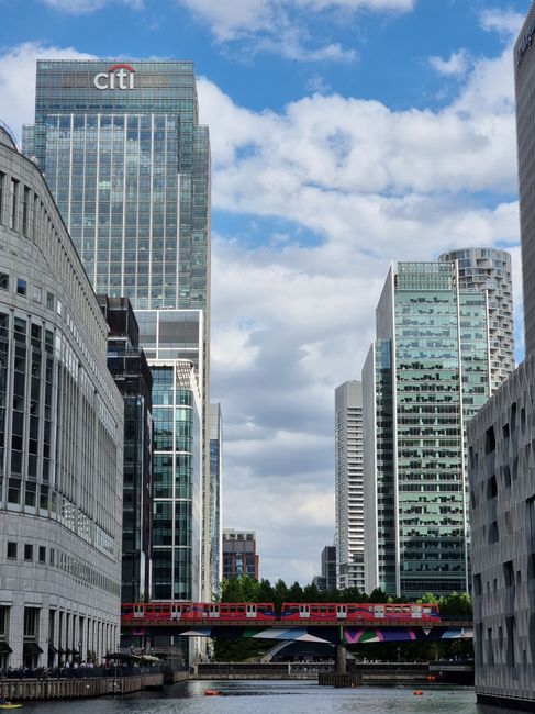 A panoramic view of Canary Wharf in London displaying modern office buildings with glass facades, including a mix of tall skyscrapers and mid-rise structures, some under construction with cranes visib